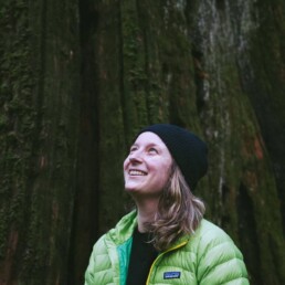 Christine Reid is smiling in front of a large tree looking up towards the sky wearing a green Patagonia jacket. Photo by Norma Ibarra